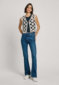 Virkat väst med blommönster i kräm över en mörk linne, matchad med blå utsvängda jeans och vita sneakers.
