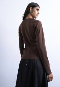 Γυναίκα με μακριά σκούρα μαλλιά που φοράει μεγάλα στρογγυλά σκουλαρίκια, καφέ μπλούζα με μακριά μανίκια και σκουρόχρωμη φούστα, κοιτώντας πάνω από τον ώμο της με φόντο ένα απλό τοίχο.