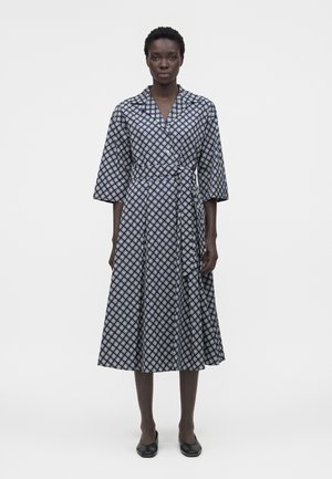 Žena stojaca v námornícky modrom midi zavinovacom šatách s bielymi geometrickými vzormi a čiernymi plochými topánkami na jednotnom pozadí.