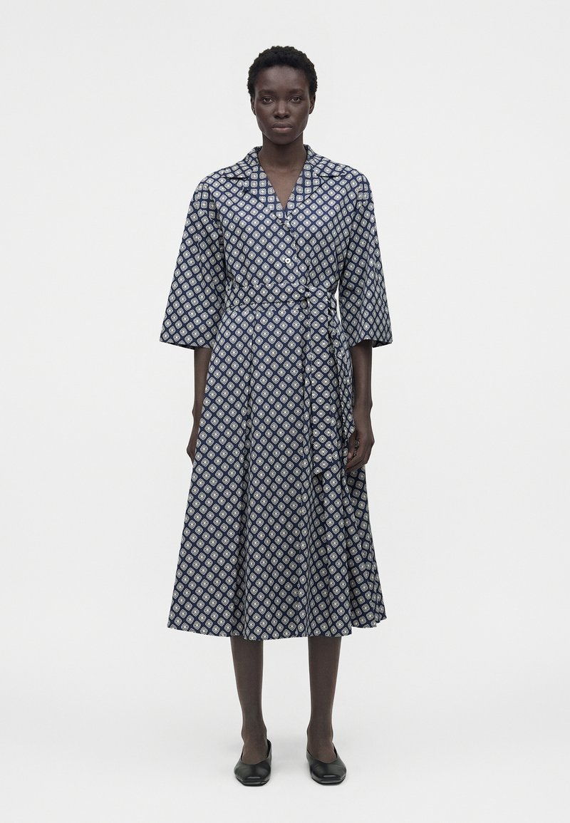 Γυναίκα που στέκεται φορώντας ένα ναυτικό μπλε μίντι φόρεμα τύπου wrap με λευκά γεωμετρικά σχέδια και μαύρα πλατιά παπούτσια, σε απλό φόντο.