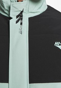 Gros plan sur une veste noire et vert clair avec le logo "1 SPORT EST. 1974" sur le rabat avant et une partie du logo "HI-TEC" sur le panneau noir.