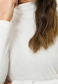 Frau mit langen braunen Haaren, die einen weißen Rollkragenpullover trägt und nachdenklich ihren Kopf auf ihre Hand stützt.