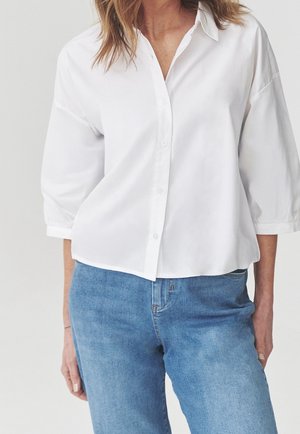 Donna che indossa una camicia bianca larga con bottoni e maniche a tre quarti e jeans in denim azzurro chiaro su uno sfondo semplice.