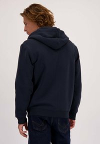 Marineblauwe hoodie met een grote capuchon, ribbelmanchetten en een zachte textuur, gecombineerd met donkere jeans. Zichtbare stiksels en achteraanzicht.