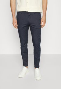 Man som bär slim-fit mörkblå byxor, vita sneakers och en ljusgul kortärmad skjorta står på ett grått golv.