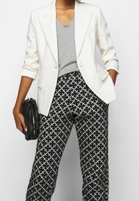 Person som bär en vit blazer, grå t-shirt, byxor med svart och vitt mönster, håller en liten svart väska med ena handen i fickan.