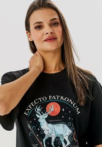 Černé tričko s grafickým designem jelena, měsíce a hvězd, s textem "EXPECTO PATRONUM" v bílé a modré barvě.