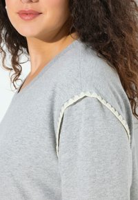 Žena s kudrnatými vlasy, oblečená v svetle šedém topu s krémovými vlnitými lemy podél ramenního švu, na bílém pozadí.