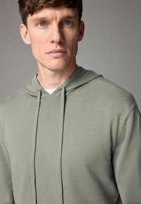Olívazöld kapucnis pulóver, puha anyagból készült. Kangaroo zsebe és hosszú zsinórjai vannak a kapucnin. Minimalista dizájn.