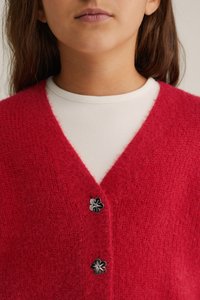 Raudona pūkuota kardiganas su V formos kaklu, puoštas juodais gėlių mygtukais ir vilkimas per baltą juostuotą marškinį.