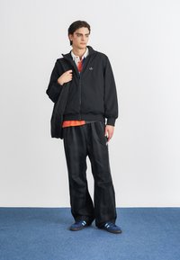 Crna bomber jakna sa zatvaračem, narančasta polo majica ispod, crne karirane široke hlače i plave tenisice s svijetloplavim prugama.