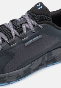 Black athletic shoe with a mesh upper, purple laces, and an Under Armour tag. Features a textured camo-patterned sole with blue accents.