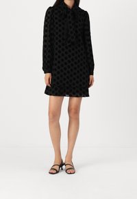 Robe noire à pois avec des manches longues, un nœud au cou et un ourlet à volants. Tissu lisse et transparent, longueur courte au-dessus du genou. Talons noirs à lanières.