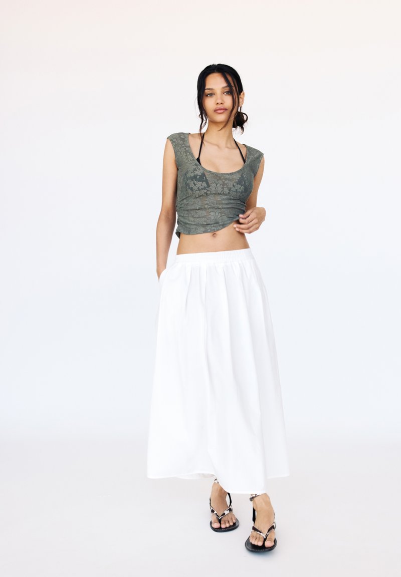 Top cropped verde oliva con stampa floreale, abbinato a una gonna bianca fluida e lunga fino alla caviglia. Il modello indossa sandali neri. Sfondo minimalista.