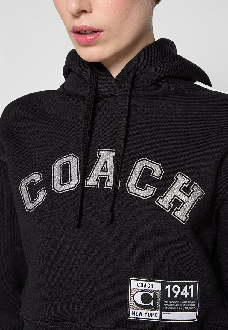 Schwarzer Hoodie mit silberner "COACH"-Aufschrift, ausgestattet mit einer Fronttasche und einem Etikett mit "COACH 1941". Strukturiertes Baumwollmaterial, Kapuze mit Kordelzug.