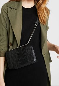 Borsa nera texturizzata con motivo a rettile, dotata di una tracolla a catena per indossarla a tracolla. Abbinata a una giacca verde oliva e un vestito nero a costine.