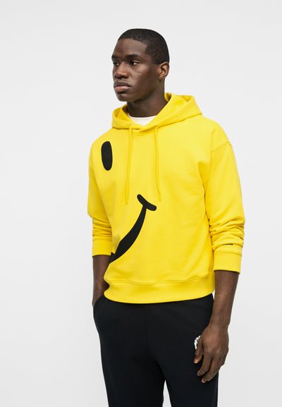 Νέος άνδρας που φοράει κίτρινο φούτερ με κουκούλα με μεγάλο μαύρο σχέδιο χαμογελαστού προσώπου, στέκεται με τα χέρια στις τσέπες μπροστά σε απλό φόντο.