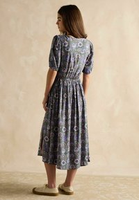 Florales Midi-Kleid mit Puffärmeln, smocktem Bund und seitlichen Schlitzen. Besteht aus grünem Stoff mit lila und blauen Mustern, kombiniert mit beigen Sneakers.