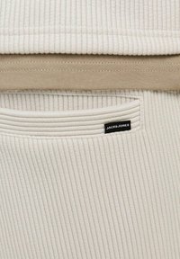 Bež ja kreem värvi sooniline kangas taskuga; funktsioonina on väike must "JACK&JONES" silt. Horisontaalne õmblusdetail loob tekstuuri.