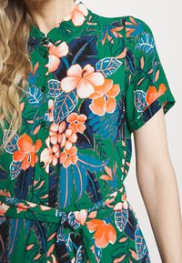Zielona sukienka w kwiatowy wzór, z krótkimi rękawami, zapinana na guziki z paskiem w talii. Posiada pomarańczowe i niebieskie kwiaty oraz akcenty liści.