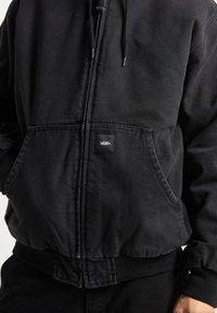 Giacca nera con cappuccio realizzata in un tessuto resistente. Presenta una zip frontale, polsini elastici e una tasca a marsupio con una patch del logo Vans.