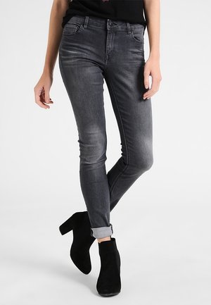 Frau trägt ausgewaschene schwarze Skinny-Jeans, an den Knöcheln hochgekrempelt, und schwarze Wildleder-Stiefeletten mit Absatz, steht mit einem Bein über das andere geschlagen.