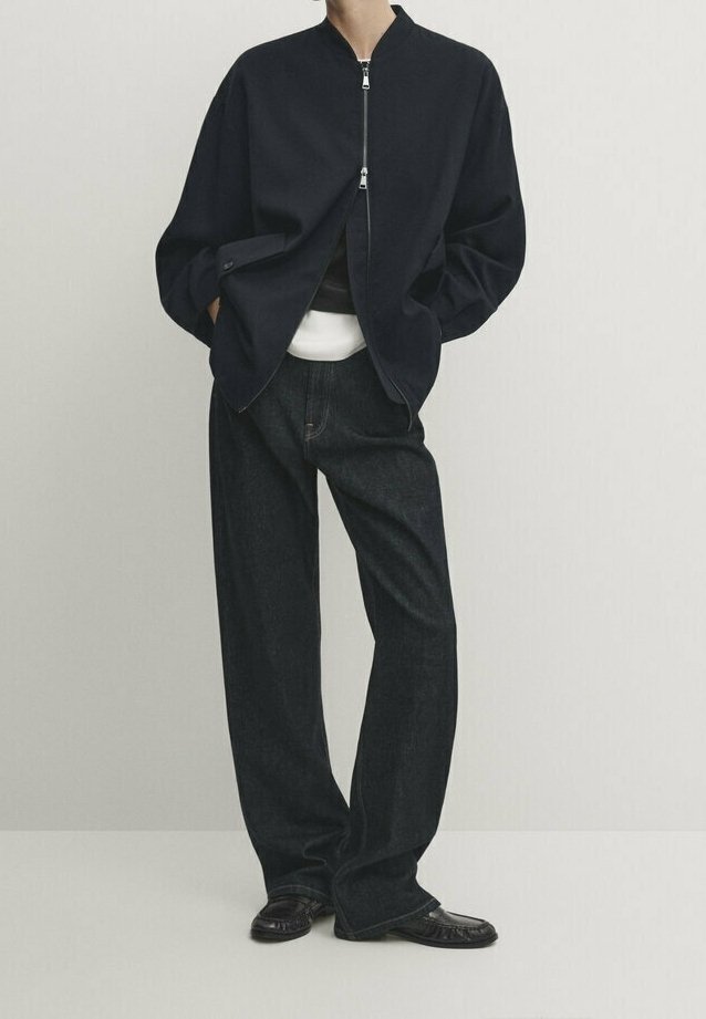 Personne portant une veste noire zippée ample, un sous-vêtement blanc, un jean large sombre et des chaussures en cuir noires, debout les mains dans les poches.