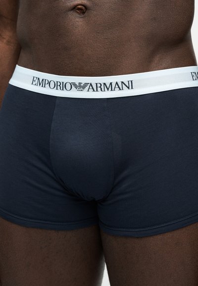 Męski tors ubrany w ciemnoniebieskie bokserki z białym paskiem w pasie z logo "Emporio Armani".