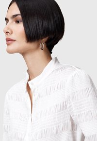 Chemise blanche texturée avec un col à boutons, ornée de plis verticaux. Accompagnée d'une boucle d'oreille en métal géométrique.