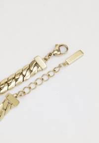 Χρυσή αλυσίδα βραχιολιού με σχέδιο υφαντής συνδέσμου, κλείσιμο με καβγά και ορθογώνια πινακίδα με χαραγμένο το "Lacoste."