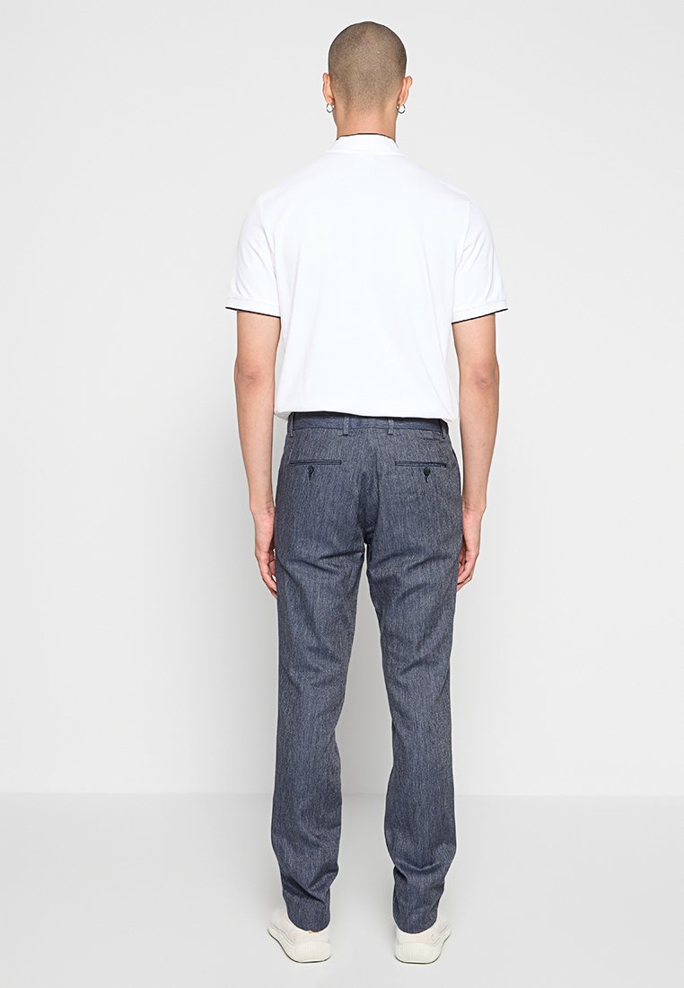Dunkelblaue Jeanshose für Herren mit geradem Schnitt, subtiler Textur und Gesäßtaschen. Kombiniert mit einem schlichten weißen Polohemd mit kurzen Ärmeln.