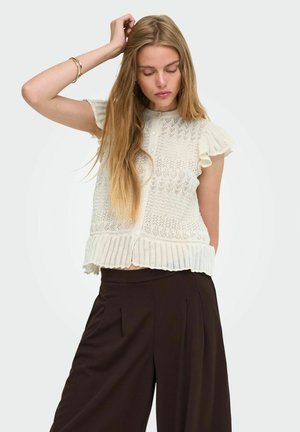 Giovane donna con lunghi capelli biondi che indossa una canotta bianca lavorata a maglia e pantaloni larghi scuri, posando davanti a uno sfondo semplice.