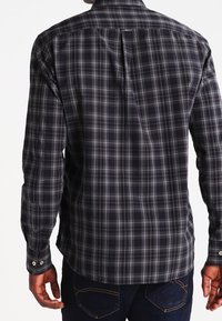 Uomo che indossa una camicia a maniche lunghe a quadri scuri e jeans scuri, visto di spalle su uno sfondo bianco.