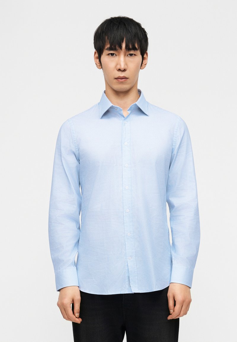 Νεαρός άνδρας που φορά ένα ανοιχτό γαλάζιο πουκάμισο με μοτίβο και μακρύ μανίκι, κουμπωτό, και μαύρο παντελόνι, στέκεται μπροστά σε απλό λευκό φόντο.