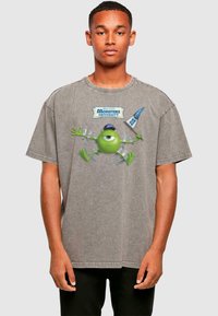 Graues, übergroßes T-Shirt mit einem Grafikdruck eines grünen Monsters mit einem Auge, umgeben von einer blauen Flagge und dem Text "Monsters University."