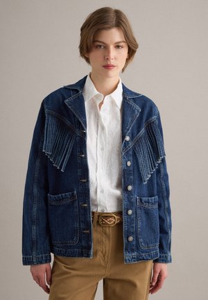 Giovane persona che indossa una giacca di jeans blu con frange, camicia bianca a fantasia, cintura marrone e pantaloni color beige su uno sfondo semplice.