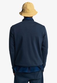Tmavě modrý pulovr s texturovaným povrchem, vybavený vysokým límcem a detailem na zadním dílu, nošený přes modrou kostkovanou košili.