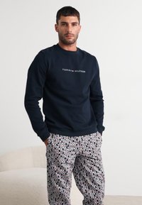 Marinegenser med hvit "TOMMY HILFIGER" logo, kombinert med mønstrede pysjamasbukser i svart, hvitt og rødt med tekst. Uformelt antrekk.