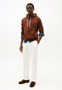 Felpa marrone con cordini bianchi, indossata sopra una camicia di jeans blu, abbinata a pantaloni bianchi e mocassini marroni. Design semplice, vestibilità rilassata.