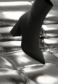 Stivaletto nero con punta appuntita e tacco a blocco appoggiato su una superficie riflettente di texture argentata. Materiale liscio con finitura opaca.