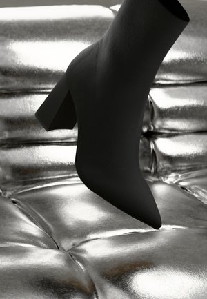Black ankle boot with a pointed toe and block heel resting above a reflective silver textured surface. Smooth material with a matte finish.