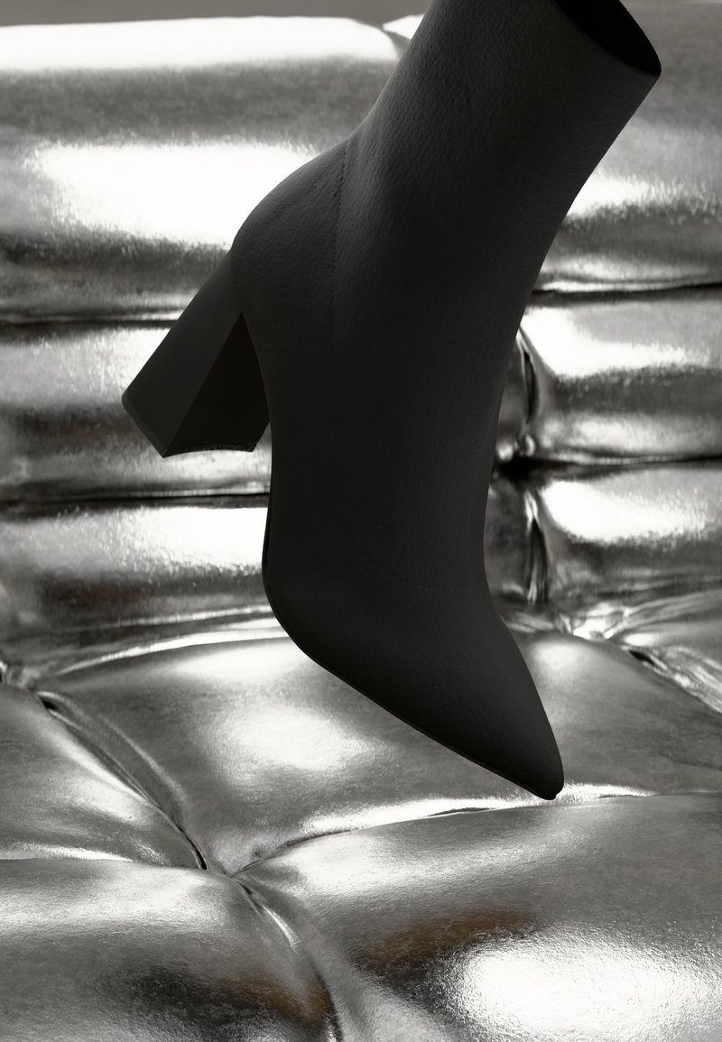 Stivaletto nero con punta appuntita e tacco a blocco appoggiato su una superficie riflettente di texture argentata. Materiale liscio con finitura opaca.