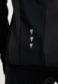 Gros plan sur une veste de sport noire avec trois triangles argentés réfléchissants orientés vers le bas sur le panneau latéral près de la taille.