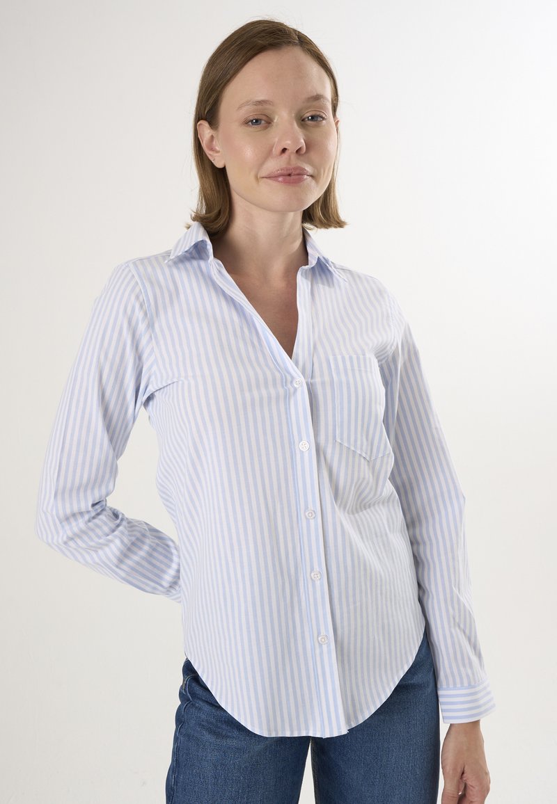 Camicia azzurro chiaro e bianca a righe con colletto, maniche lunghe e tasca sul petto, indossata con jeans scuri. Tessuto dalla texture liscia.