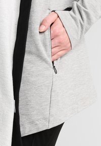 Material de sudadera gris con un borde negro, presenta un bolsillo lateral con cremallera y una textura suave, diseñado para la comodidad y la funcionalidad.