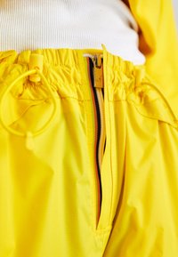 Pantaloni impermeabili gialli con vita elastica, cordino e cerniera con una striscia in contrasto. Texture liscia con una finitura lucida.