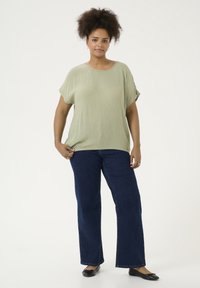 Helles grünes, weit geschnittenes Blusenshirt mit kurzen Ärmeln; kombiniert mit dunkelblauen, weit geschnittenen Jeans und schwarzen flachen Schuhen. Weißer Hintergrund.
