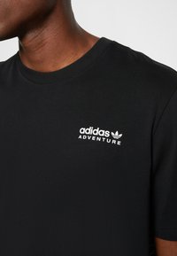 T-shirt nera in cotone con collo rotondo e maniche corte. Logo bianco "adidas ADVENTURE" sul petto a sinistra. Tessuto liscio.