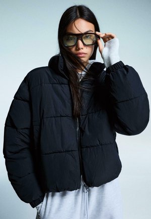 Black puffy jacket with a zip front, elastic cuffs, and quilted texture. Model wears grey sweatshirt and oversized sunglasses.