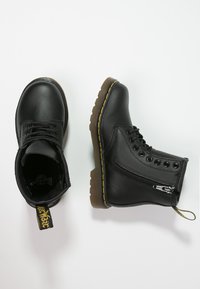 Botas de cuero negro hasta el tobillo con cordones en la parte frontal, cremallera lateral y costuras amarillas en la suela de goma. Parte superior texturizada y etiqueta en el talón.